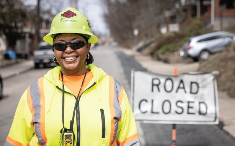 female team member in construction yellow smiling in front of a road closed sign
