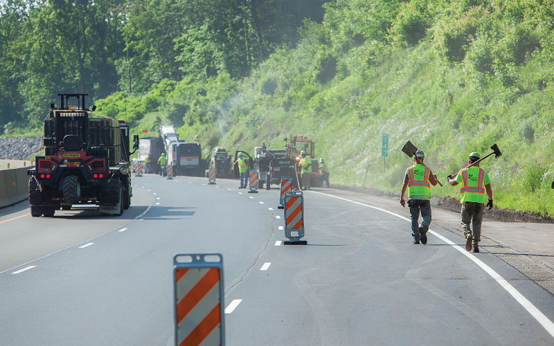 two worker carrying shovels walking along the highway towards their team working machinery to clear the shoulder of debris